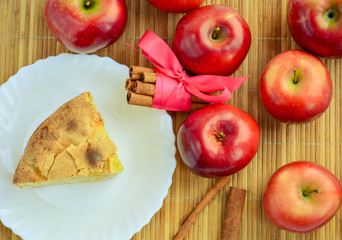 Appetizing piece of apple pie on a white plate next to cinnamon and red apples