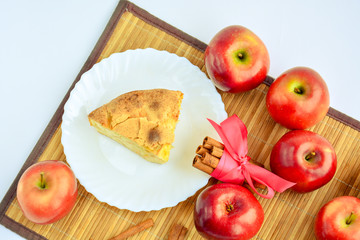 Appetizing piece of apple pie on a white plate next to cinnamon and red apples