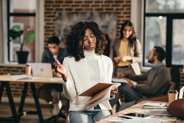 focused african american casual businesswoman holding journal with colleagues working behind in loft office