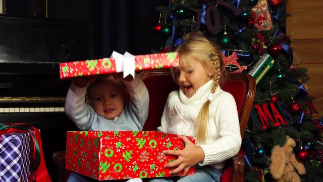 Excited siblings open up big surprise wrapped gift box with presents from Santa under Christmas tree on New Year holiday