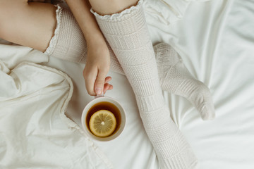 Obraz na płótnie Canvas Cozy flatlay of woman's legs in warm white stockings in bed holding cup of lemon tea, selective focus