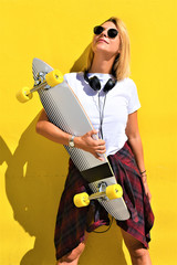 Portrait of happy beautiful blonde girl hold her wooden longboard skateboard on yellow background wall, look to the camera and smile. Urban scene, city life. Hipster cute lady. © bobanphotomkd