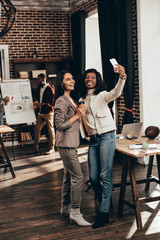 smiling multiethnic couple of business women taking selfie on smartphone in loft office with colleagues on background