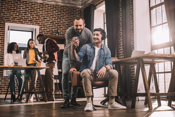 two men using smartphone with multiethnic group of casual businesspeople working in modern loft office behind