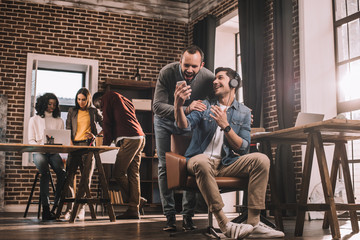 Fototapeta premium two smiling men using smartphone with multiethnic group of casual businesspeople in modern loft office behind
