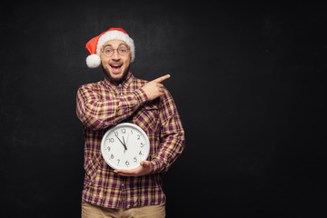 Christmas men with clock. Portrait of surprised young man wearing red santa claus hat, holding clock in hands, on black background. Fun emotion facial expression. Last minute christmas shopping