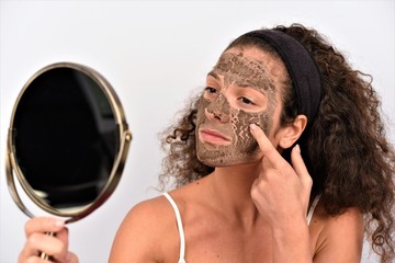 Woman removing facial dried clay mud mask in front of mirror. Skin care. Girl taking care of her complexion. Beauty spa treatment.