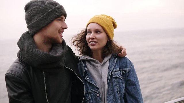 Hipster young happy couple standing together and hugging at the seaside. Young people wearing warm clothes, hats in cold weather.Looking to each other, smiling, talking. Close up