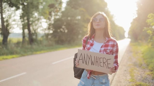 Young beautiful woman hitchhiking standing on the road holding anywhere sign. Summer time