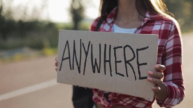 Young beautiful woman hitchhiking standing on the road holding anywhere sign. Summer time