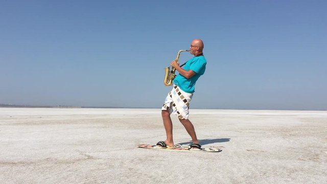  Saxophonist plays and stands on the board for a kite against the background of white salt lake