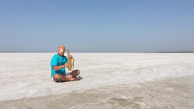 The Saxophonist Sits In The Lotus Position And Plays The Saxophone On The Background Of White Salt Desert Moove Camera