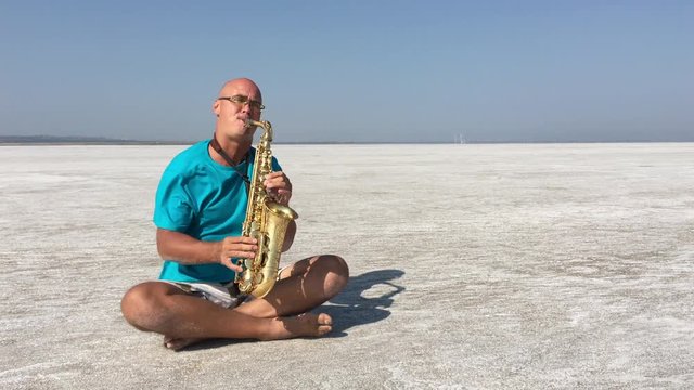 The Saxophonist Sits In The Lotus Position And Plays The Saxophone On The Background Of White Salt Desert