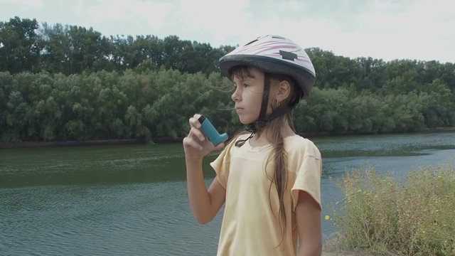 A child with asthma. A cute little girl in a bicycle helmet uses an inhaler outdoors. Child's asmatic attack in nature.