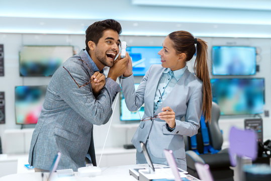 Multicultural couple dressed in business clothes trying out 360 degree camera while standing in tech store. In background plasma television.