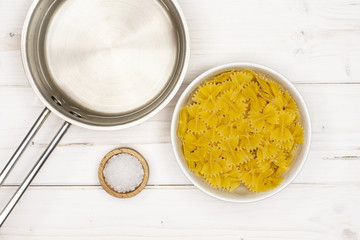 Raw farfalle with water in stewpan and salt. recipe step by step farfalle with arugula leaves flatlay on white wood