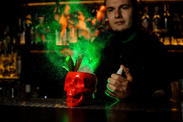 Barman sprinkling bitter into a glass in shape of red bone scull filled with drink decorated with...