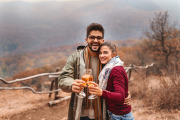 Fototapeta na wymiar Happy couple hugging and making a toast outdoors while looking at camera. Autumn season. Forest and mountains in background.