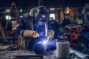 Worker welding iron. Protective suit and mask on. Workshop interior.