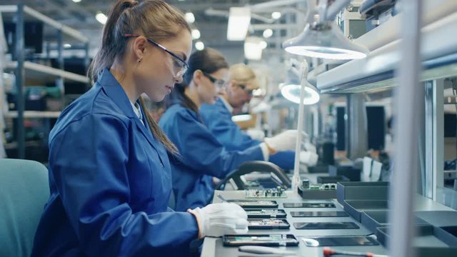 Female Electronics Factory Worker in Blue Work Coat and Protective Glasses is Assembling Printed Circuit Boards for Smartphones with Tweezers. High Tech Factory with more Employees in the Background. 