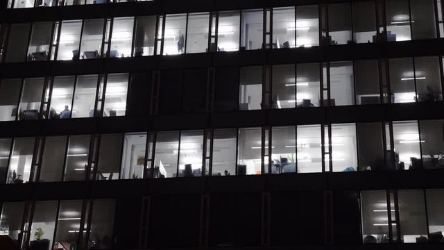 Modern multistory building facade, office center at dusk, windows with electric lighting and silhouettes of working people inside, side view, stock video
