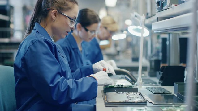 Female Electronics Factory Worker in Blue Work Coat and Protective Glasses is Assembling Laptop's Motherboard with a Screwdriver