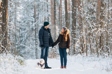 Fototapeta na wymiar Cute young couple having fun in winter forest with their pretty little white dog. Man and woman in sweater with a snowflake. Christmas and winter holidays