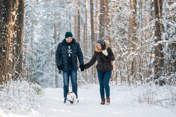 Cute young couple having fun in winter forest with their pretty little white dog. Man and woman in sweater with a snowflake. Christmas and winter holidays