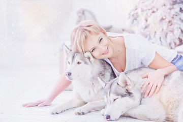 Beautiful blonde woman sits with two husky dogs near christmas tree.