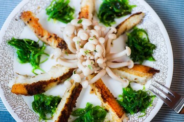 Fried fish, shimeji mushroom and seaweed on plate with fork.