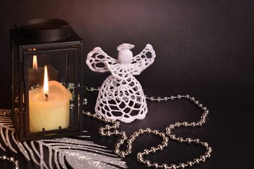 Christmas decoration on black background. White crochet angel, lantern, candle, silver feather and beads