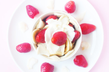 Healthy raw vegan dessert - banana and strawberry with coconut cream and coconut chips on pink background