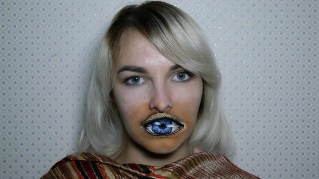 Pretty young girl with long blonde hair watch forward with big blue painted eye on her lips. Strange lady with frick makeup in room. Concept body art on face with beautiful model . Wink with eye 
