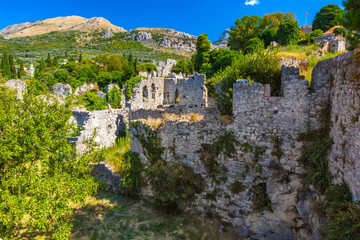 Fototapeta na wymiar The picturesque historic walls of the destroyed city covered with green plants grass trees on the backdrop of colorful mountains and clear sky. Summer landscape in Fortress Old Bar Town, Montenegro.