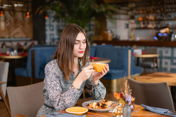 Young woman drinking coffee. Female holding cup. Sitting in coffee shop at wooden table. On table plate with breakfast. pleasant morning, the beginning of the working day