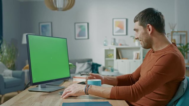 Handsome Man Sitting at His Desk at Home Uses Personal Computer with Mock-up Green Screen. He Drinks Beverage from the Mug.
