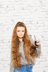 Fototapeta na wymiar Teenage girl with long blonde hair, in casual outfit, holding a glass with cola drink, standing against brick wall. Studio lighting, no retouch.