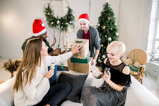 Picture showing group of friends with Christmas presents, celebrating Christmas or New Year in cozy decorated living room. Two boys, African and Caucasian in santa hats with two pretty Caucasian girls
