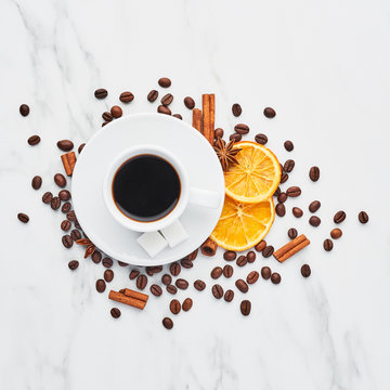 Cup of coffee with beans, chocolate, dried oranges, anis and cinnamon sticks on white marble background. Concept of coffee with different spices. Top view, square crop.