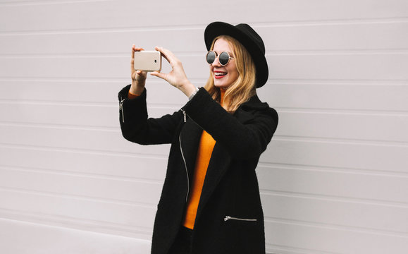 Fashion, technology and people concept - stylish smiling woman taking selfie picture by smartphone, elegant female model in black jacket coat, round hat in city on gray wall background