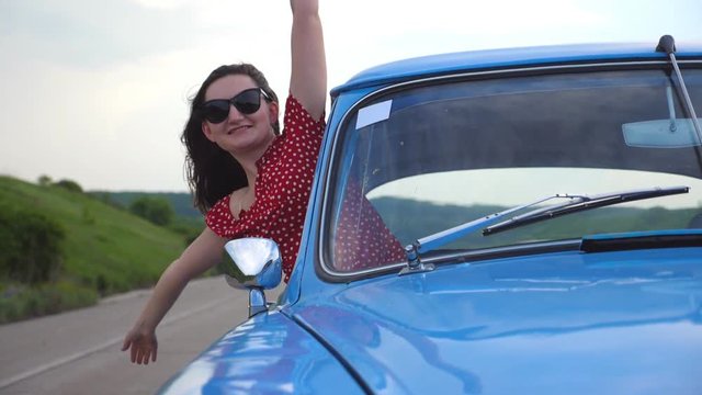 Young smiling girl in sunglasses leaning out of vintage car window and enjoying trip. Happy attractive woman looks out from moving retro auto and raising hands. Travel and freedom concept. Slow motion