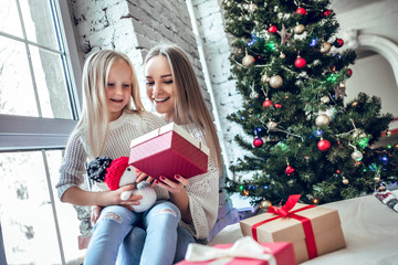 Happy mother and daughter with gifts near Christmas tree are smiling and holding the gift box.