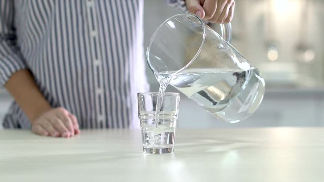 Woman pouring water from jug into glass on table indoors, closeup
