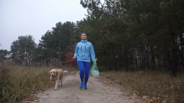 Environment friendly sporty woman with her labrador retriever running with trash bags doing plogging in autumn woodland during morning jog. Positive fitness female runner with pet plogging in forest.