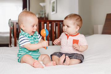 Group portrait of two white Caucasian cute funny baby boys sitting together on bed sharing toy....