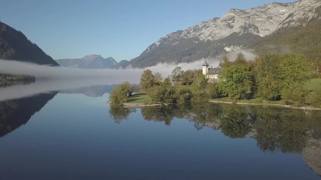 Aerial, drone shot around a church, on a bay, mountains reflecting from a lake, on a sunny and foggy morning, at Grundlsee, in Liezen, Austria