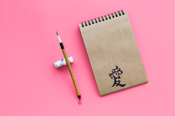 Chinese traditional calligraphy concept. Asian hieroglyph love in craft paper notebook near special writting pen on pink background top view copy space