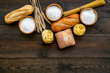 Farinaceous food. Fresh bread and raw pasta near flour in bowl and wheat ears on dark wooden background top view space for text