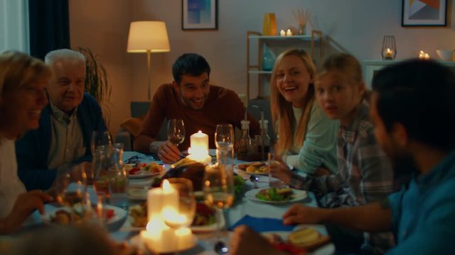 In the Evening: Family and Friends Gathered at the Dinner Table. Old and Young People Have Fun, Eating, Drinking. Cozy Living Room Atmosphere with Warm Candle Light Illumination.