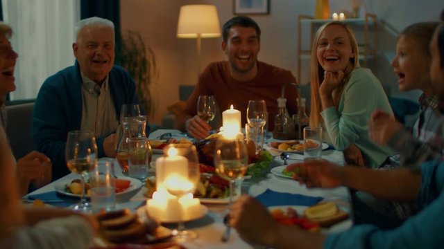 In the Evening: Family and Friends Gathered at the Dinner Table. Old and Young People Have Fun, Eating, Drinking. Applaud and Propose a Toast. Cozy Living Room Atmosphere.
